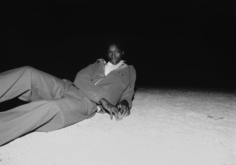 A dark-skinned woman reclines on light grey pavement in a grey jumpsuit with a white top against a dark, almost black background.