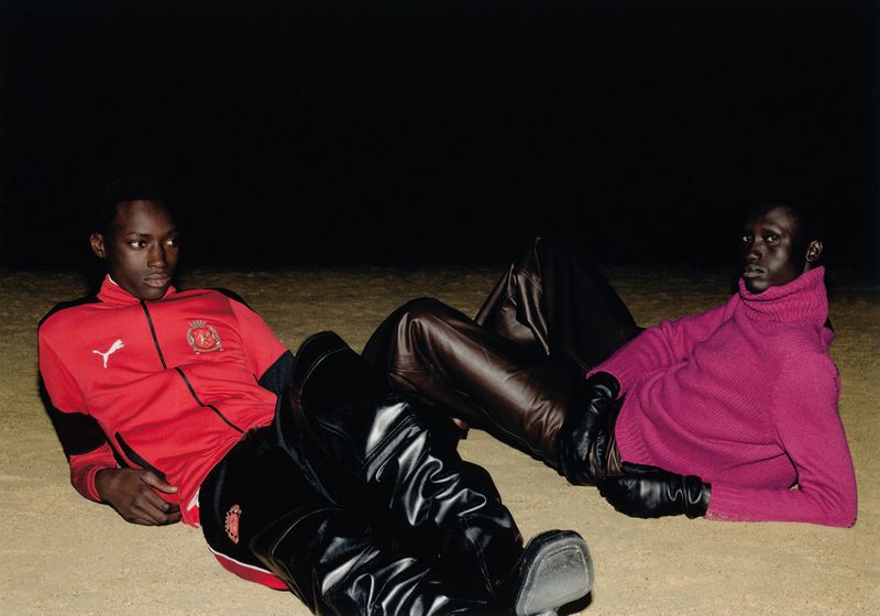 Two dark-skinned male fashion models lying on the ground in a dimly lit environment wearing tracksuits and leather pants and gloves.