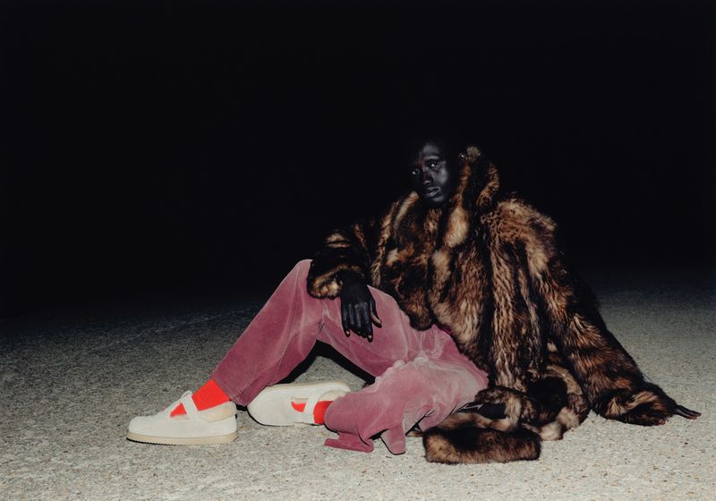 Person wearing a large fur coat, pink pants, and cream sneakers sitting in a dimly lit environment with a dark background.
