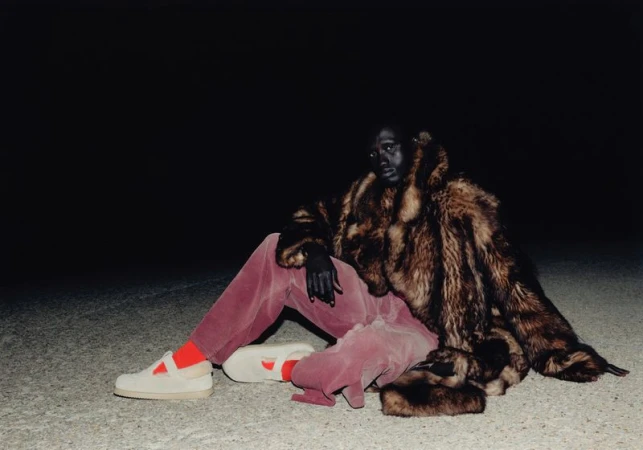 Person wearing a large fur coat, pink pants, and cream sneakers sitting in a dimly lit environment with a dark background.