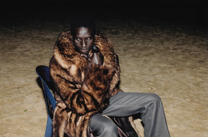 A dark-skinned person sits on a blue chair in a fur coat and grey pants in a dimly lit outdoor setting, looking down with a serious expression.