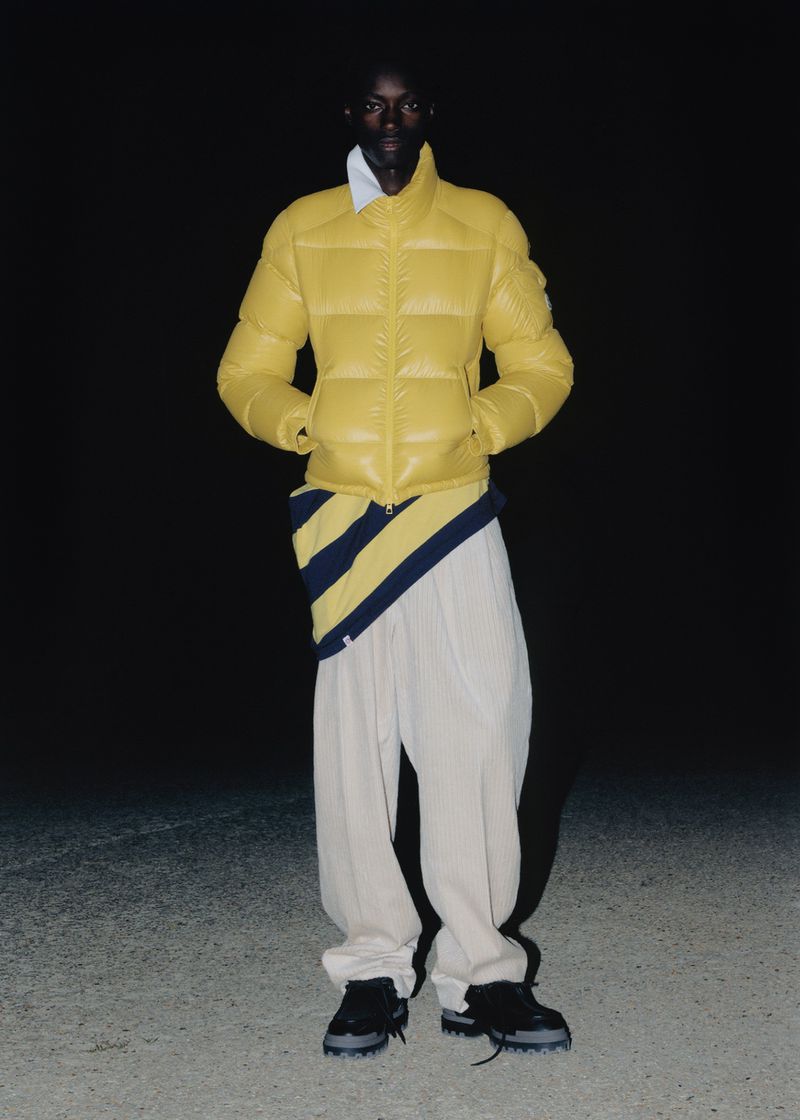 A dark-skinned male fashion model is standing, wearing a yellow puffer jacket, striped shirt, white pants and black shoes against a dark background.