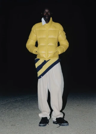 A dark-skinned male fashion model is standing, wearing a yellow puffer jacket, striped shirt, white pants and black shoes against a dark background.