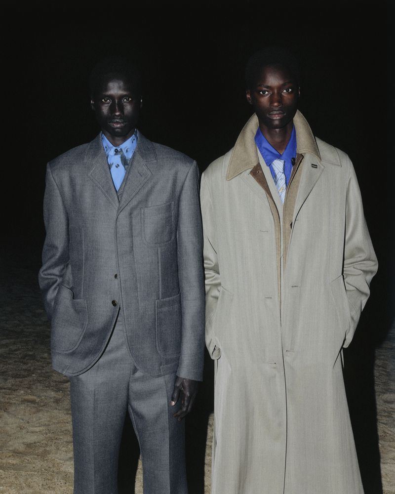 Two adult males in formalwear stand side-by-side against a dark backdrop. One wears a grey suit, the other a tan trench coat.