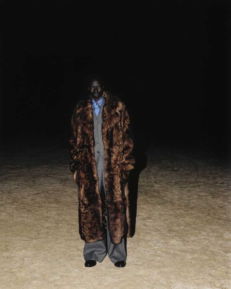 Solo male fashion model wearing a long fur coat and grey suit standing in an outdoor environment at night with dark background.