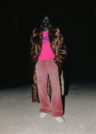 Black man posing outdoors at night wearing a long brown fur coat, hot pink graphic t-shirt, and wide-leg pink corduroy trousers.