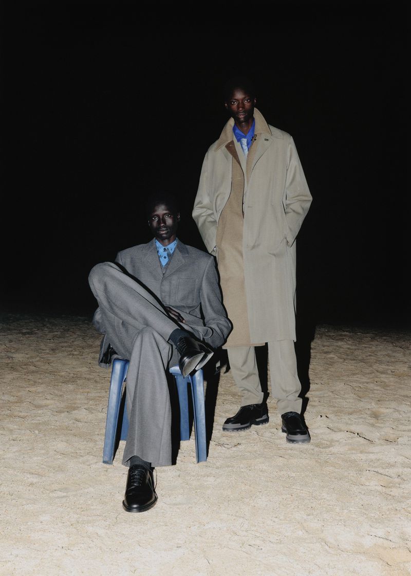 Two adult men pose in dark outdoor setting: one seated in grey suit on plastic chair, the other standing in a tan trench coat and black shoes.