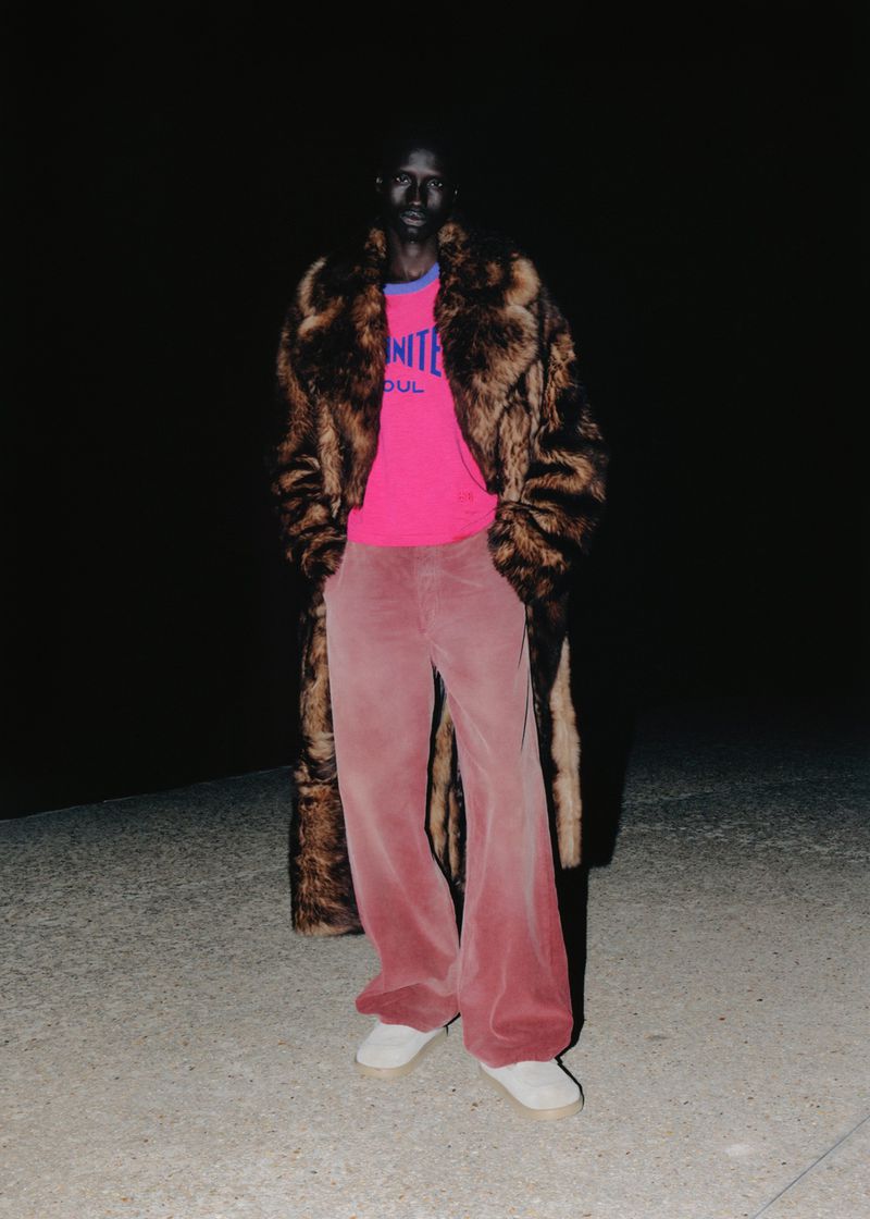 Black man posing outdoors at night wearing a long brown fur coat, hot pink graphic t-shirt, and wide-leg pink corduroy trousers.