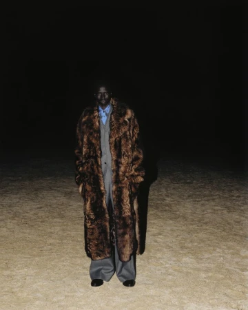 Model wearing a full-length brown fur coat over a grey suit standing on sand against a pitch black night background.