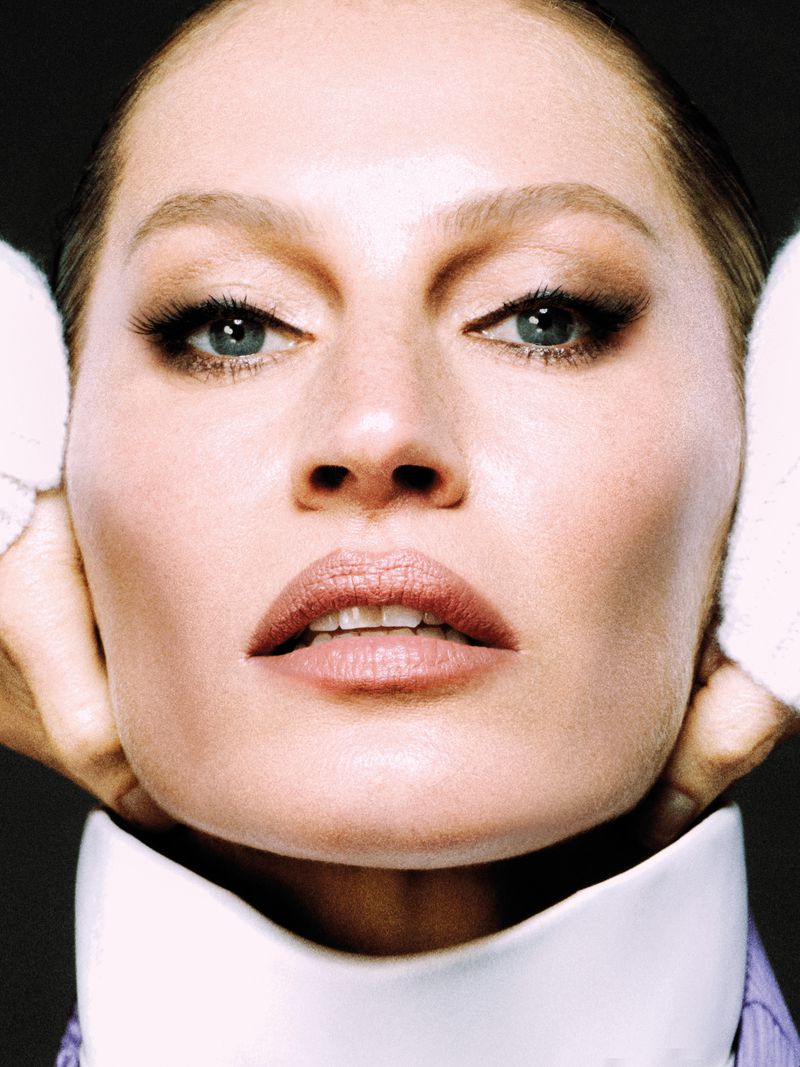 Close-up portrait of a woman with makeup and a white collared shirt, hands gently framing her face, set against a dark gray backdrop.