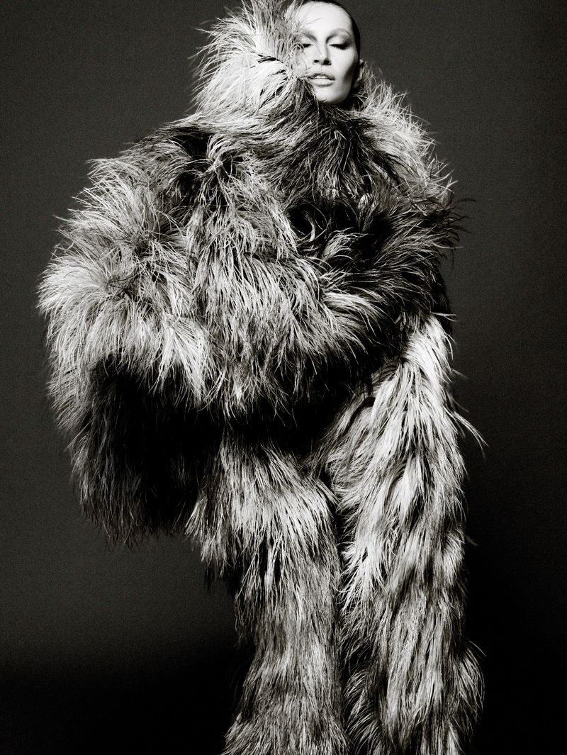 Black and white portrait of a person in a large feathered coat, featuring dramatic lighting and a neutral studio background.