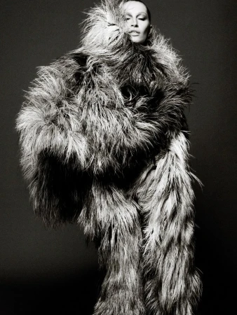 Black and white portrait of a person in a large feathered coat, featuring dramatic lighting and a neutral studio background.