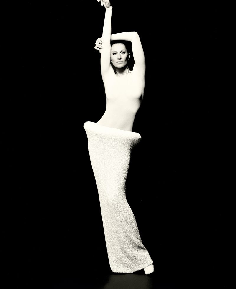 Elegant adult female in flowing white gown poses in a studio portrait with dark backdrop, arms raised, showing fashion and artistry.