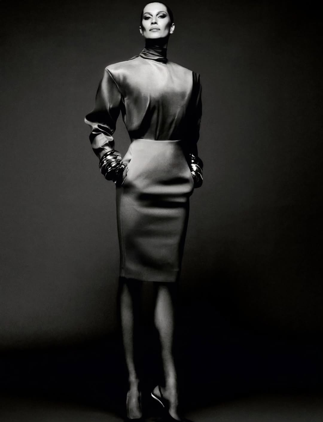 Elegant full-length portrait of an adult woman in a satin high-neck blouse, pencil skirt, and chunky jewelry against a gray backdrop.
