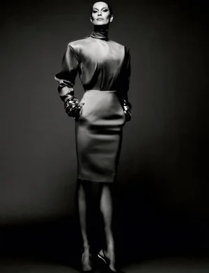 Gisele Bündchen posing in a high-neck blouse, pencil skirt, and stacked metallic bangles against a dark studio background.