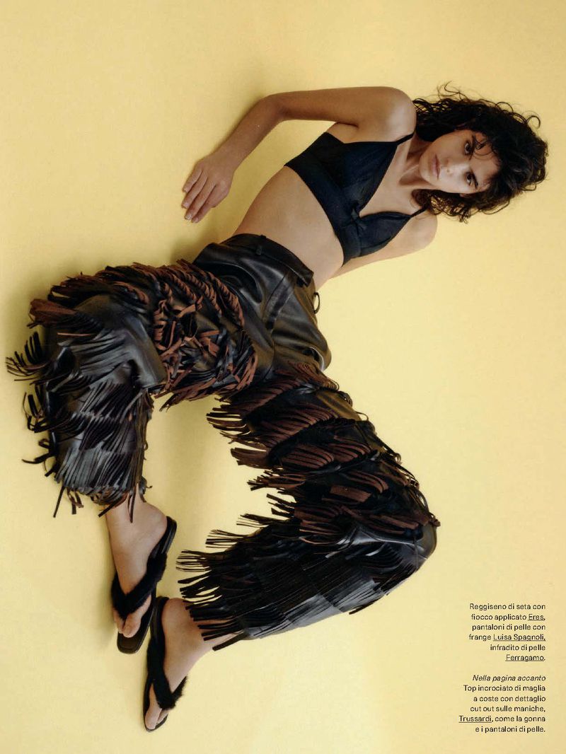 A slender woman with curly dark hair poses in a studio. She wears a black bra top, fringed leather pants, and black flip-flops against yellow backdrop.