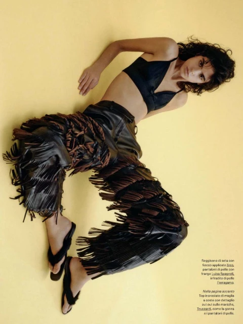 A slender woman with curly dark hair poses in a studio. She wears a black bra top, fringed leather pants, and black flip-flops against yellow backdrop.