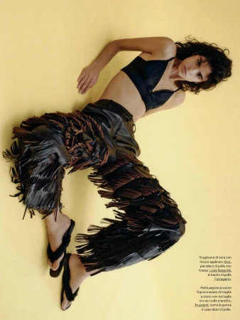 A slender woman with curly dark hair poses in a studio. She wears a black bra top, fringed leather pants, and black flip-flops against yellow backdrop.