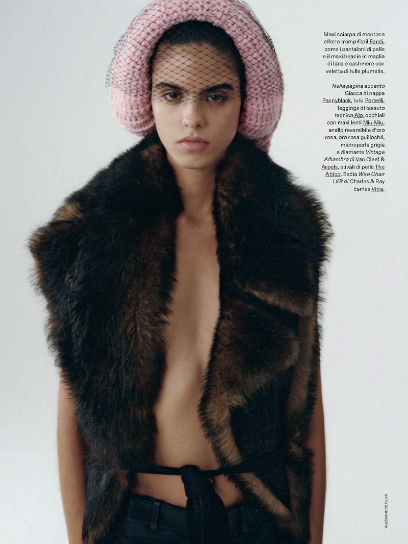 A young, serious-looking woman poses in a studio with a fur vest and a pink hat with a dark veil over her face in a medium shot.