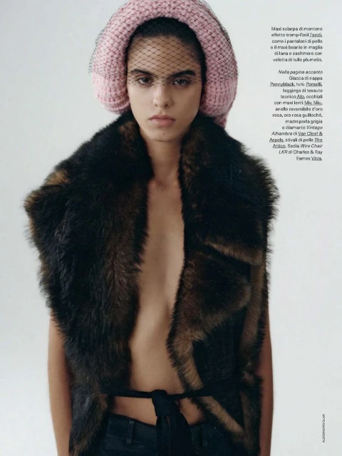 A young, serious-looking woman poses in a studio with a fur vest and a pink hat with a dark veil over her face in a medium shot.