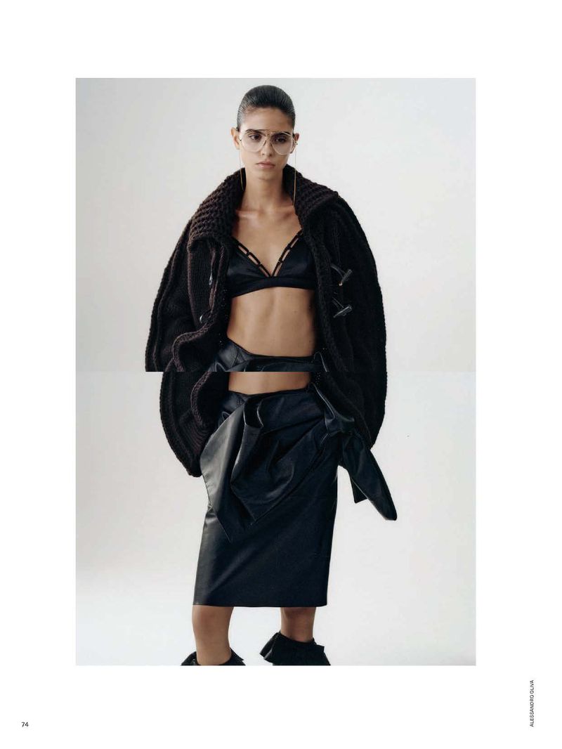 Fashion editorial shot of a woman in a black bralette, skirt, and coat posing in a studio with soft, diffused lighting and a neutral background.
