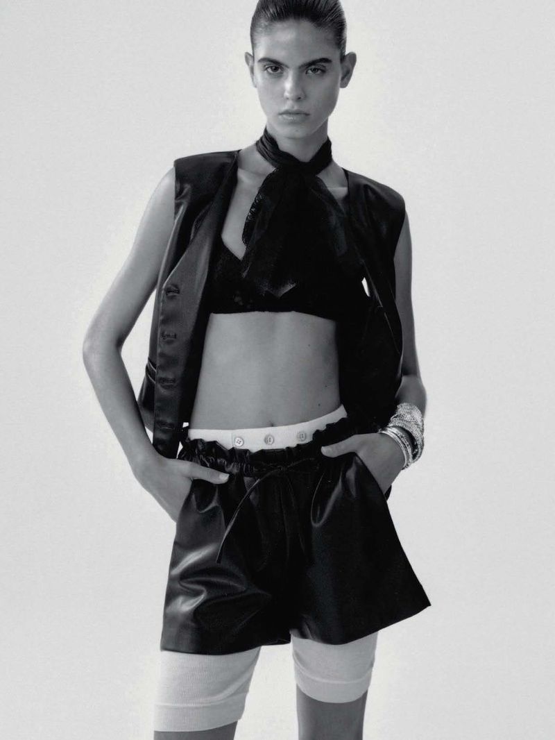 Fashion shot of a slender woman wearing a black vest, bra, shorts, white socks, and silver bracelets in front of a neutral background.