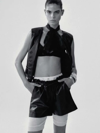 Fashion shot of a slender woman wearing a black vest, bra, shorts, white socks, and silver bracelets in front of a neutral background.