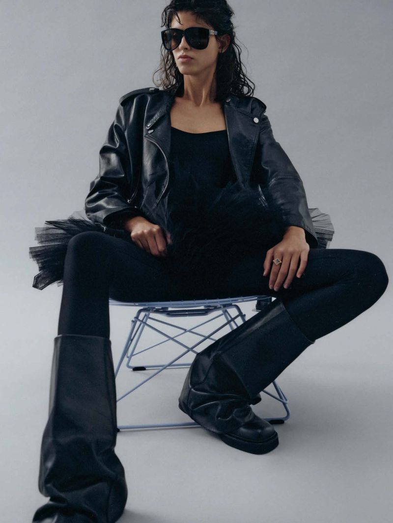 Adult female in black leather jacket, tank top, tights, and boots sitting on a transparent chair in a photo studio against grey backdrop.