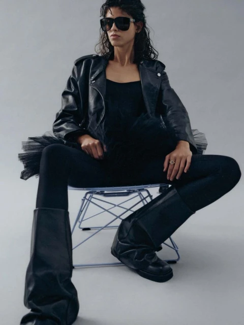 Adult female in black leather jacket, tank top, tights, and boots sitting on a transparent chair in a photo studio against grey backdrop.