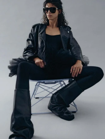 Adult female in black leather jacket, tank top, tights, and boots sitting on a transparent chair in a photo studio against grey backdrop.