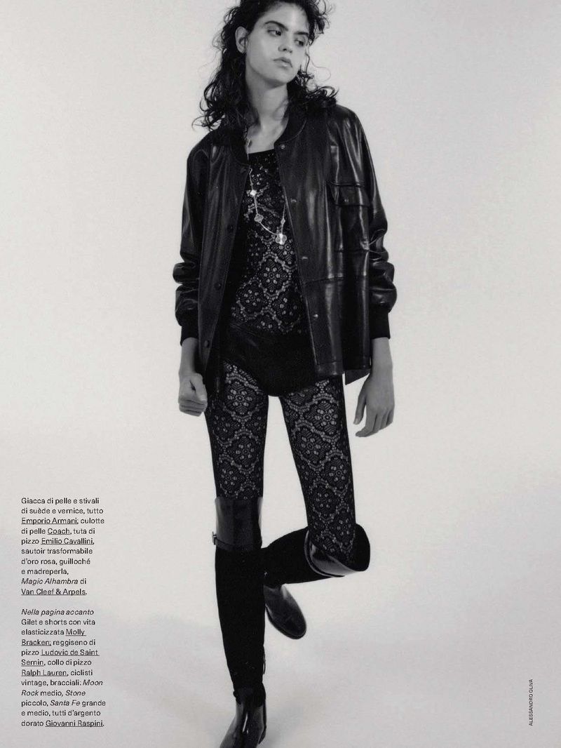 A young woman in black leather jacket, lace top and tights poses in a studio with a white background.