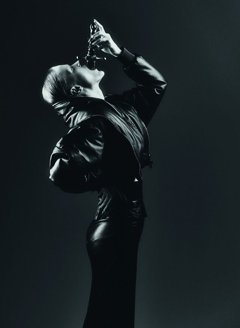 A woman in a black leather jacket and skirt drinks from a black bottle in a dark studio, captured in an artistic black and white image.