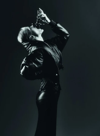 A woman in a black leather jacket and skirt drinks from a black bottle in a dark studio, captured in an artistic black and white image.