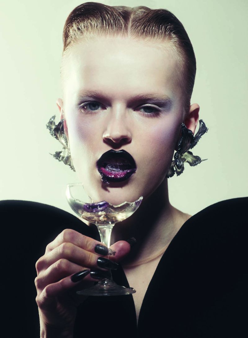 A stylish portrait of a woman with black lipstick holding a shattered glass, wearing large earrings, and posing in front of a pale background.