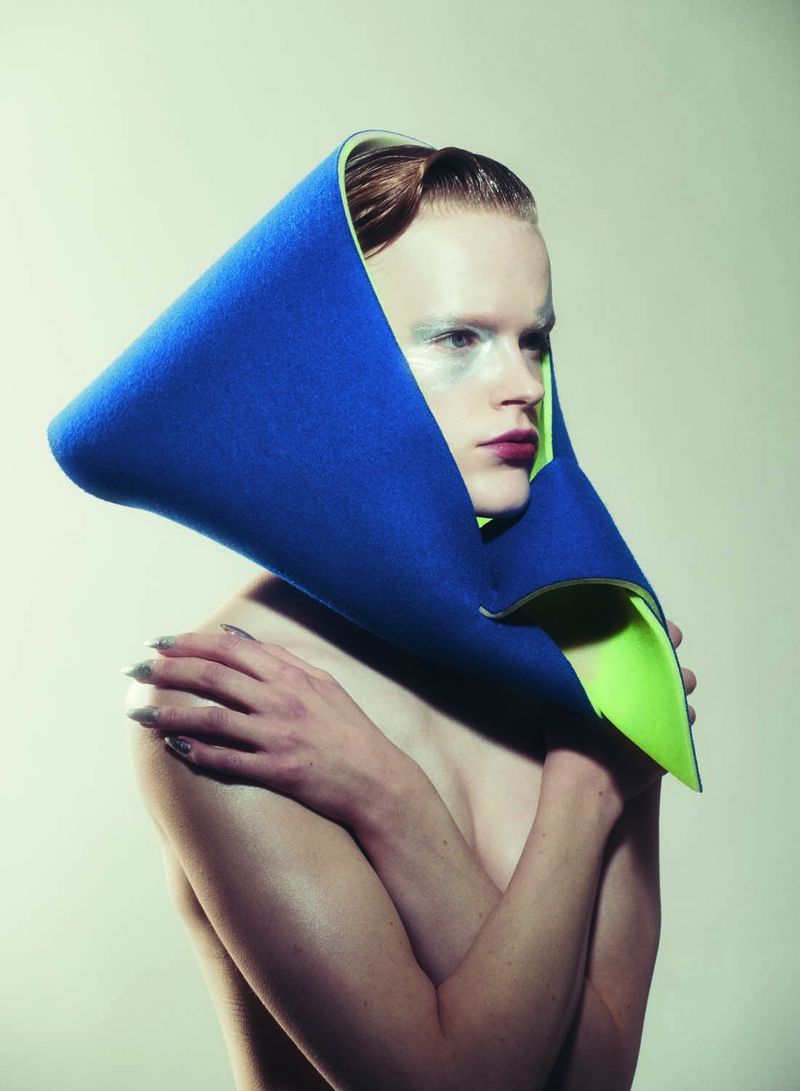 Headshot of androgynous fashion model with dramatic makeup, blue and neon green collar, and silver fingernails posing in photo studio.