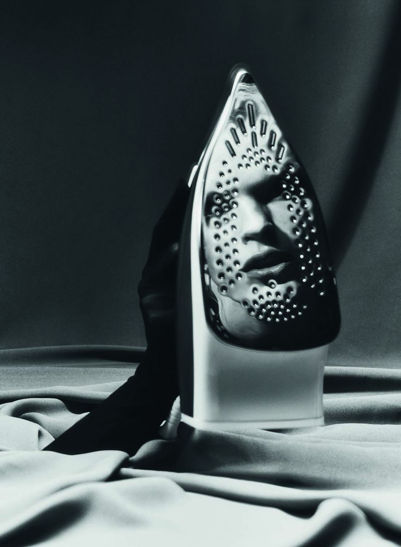 Monochrome studio shot of an iron reflecting a human face, set against a draped fabric backdrop. The light and shadow create a surreal effect.