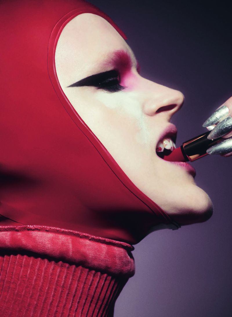 Adult female model applies red lipstick, wearing a matching red turtleneck and hood, along with dramatic black and pink eyeshadow.