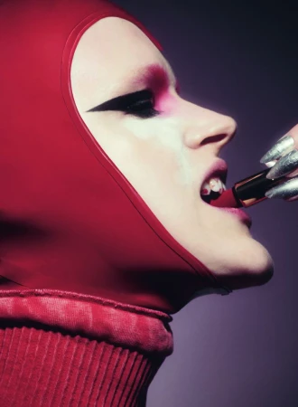 Adult female model applies red lipstick, wearing a matching red turtleneck and hood, along with dramatic black and pink eyeshadow.