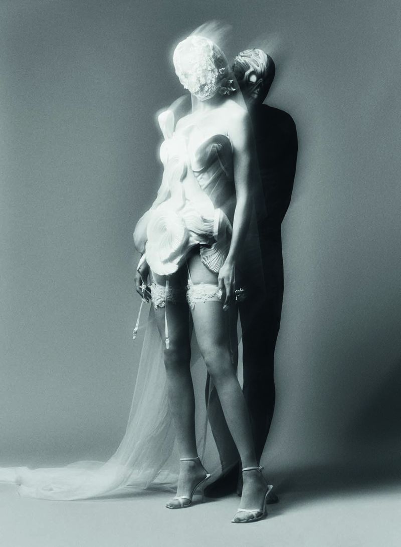 Blurred motion studio shot of a couple. The woman wears a corset dress, gartered stockings, and heels while being embraced by a male figure.