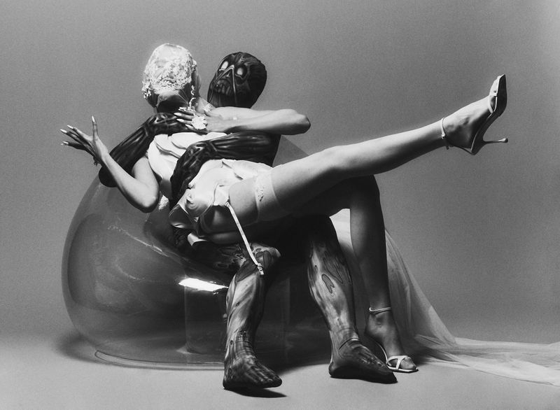 Two figures, one in an elaborate white headpiece and lingerie, the other in a dark, abstract costume, embrace in a clear chair.