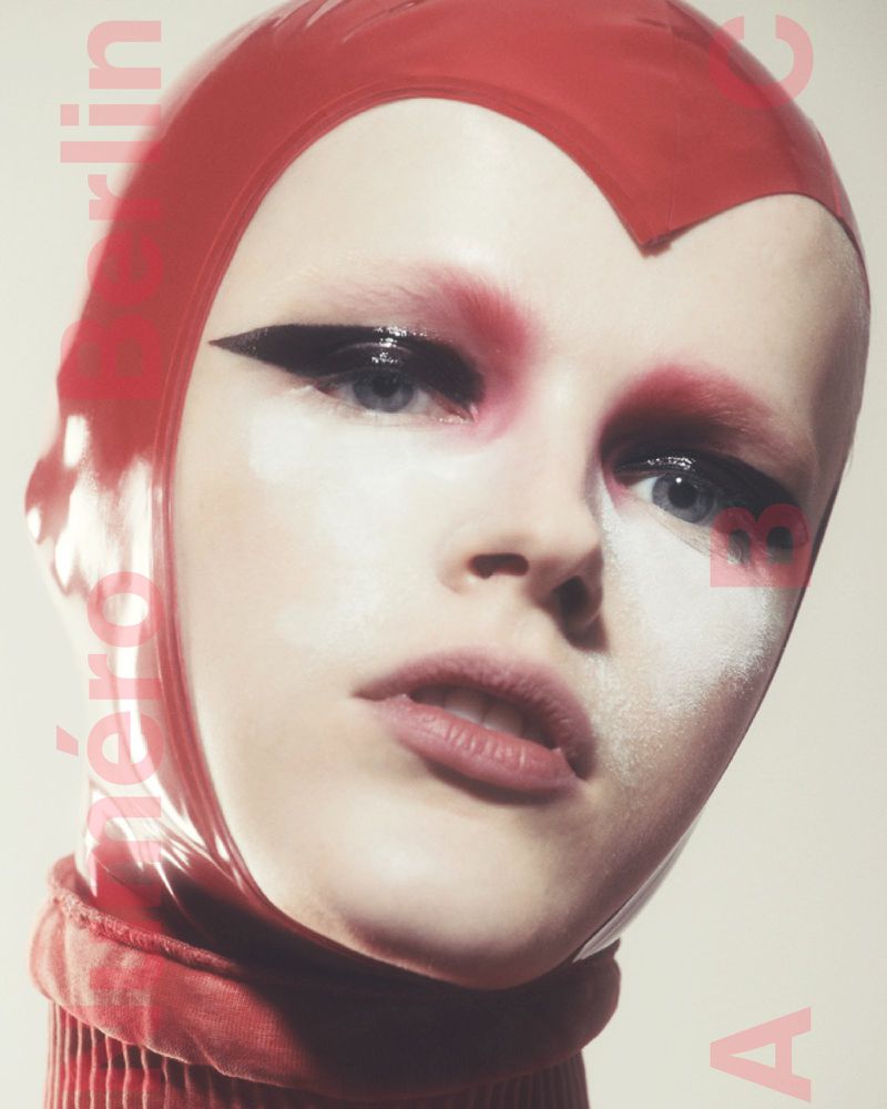 Striking close-up studio portrait of a fair-skinned person wearing a red latex hood with stylized makeup and a high collar.