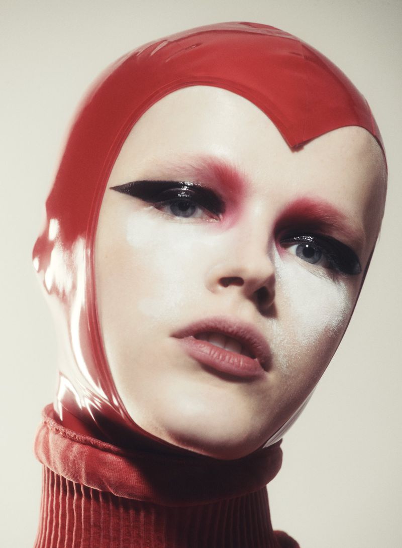 Headshot of a person with dramatic makeup and a red hood against an off-white background in a beauty or fashion editorial-style image.
