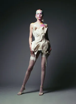 Model Hannah Motler wearing a sculptural white corset and lace stockings against a grey studio backdrop.