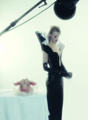 Model in black avant-garde outfit with high collar standing next to a table with a pig head for Numéro Berlin Beauty