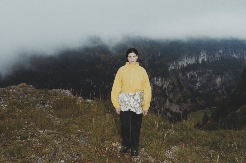 A woman in a yellow top and dark pants stands on a grassy hilltop with mountains and a cloudy sky in the distance. Lifestyle shot.