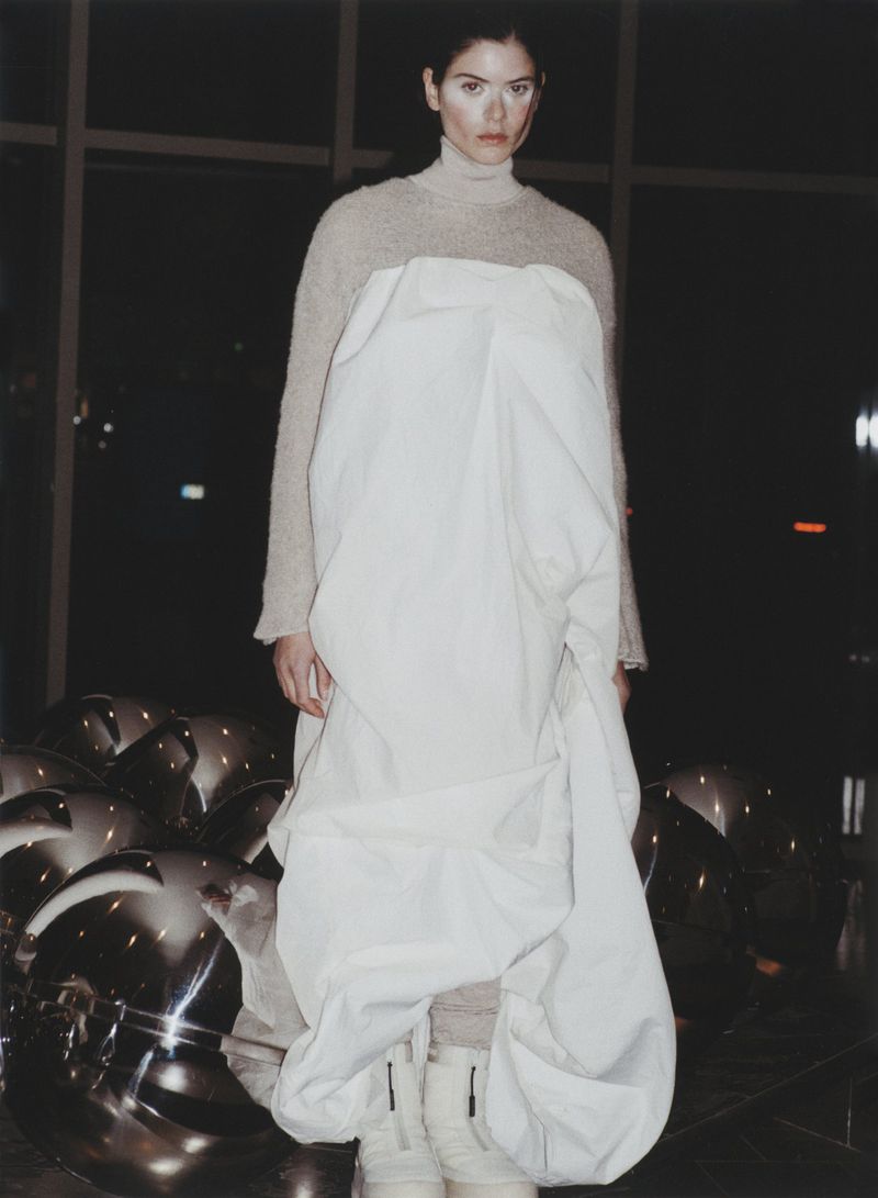 A solitary woman in a draped white dress stands against a dark backdrop with mirrored spheres, creating an ethereal and reflective fashion image.