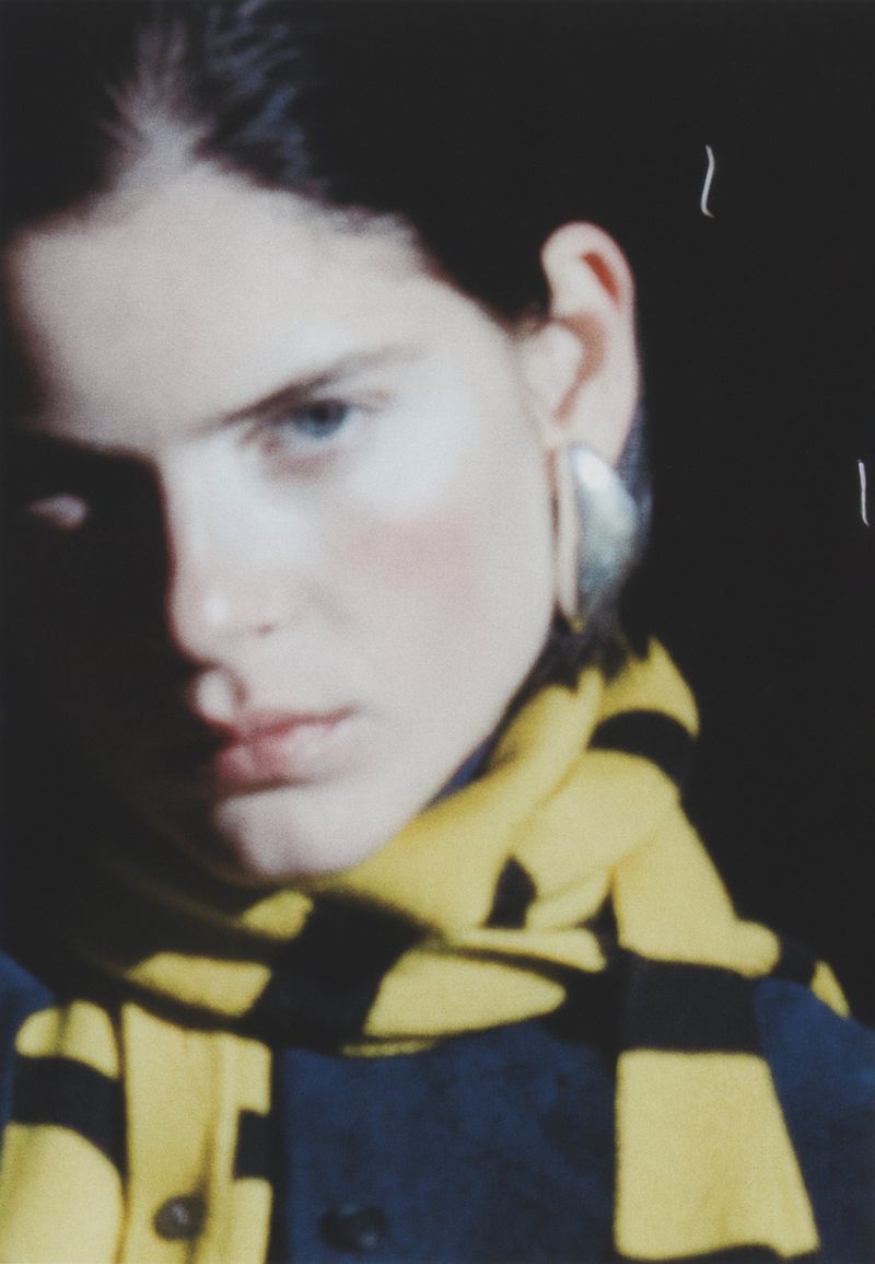 Close-up studio portrait of a young adult woman with blue eyes and fair skin wearing statement earrings and a yellow and black striped scarf.