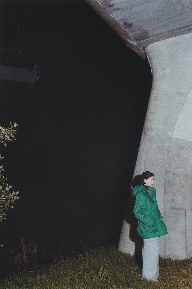 Woman in green coat stands beneath a concrete overpass at night. Dark background with white flowering tree and green grasses.