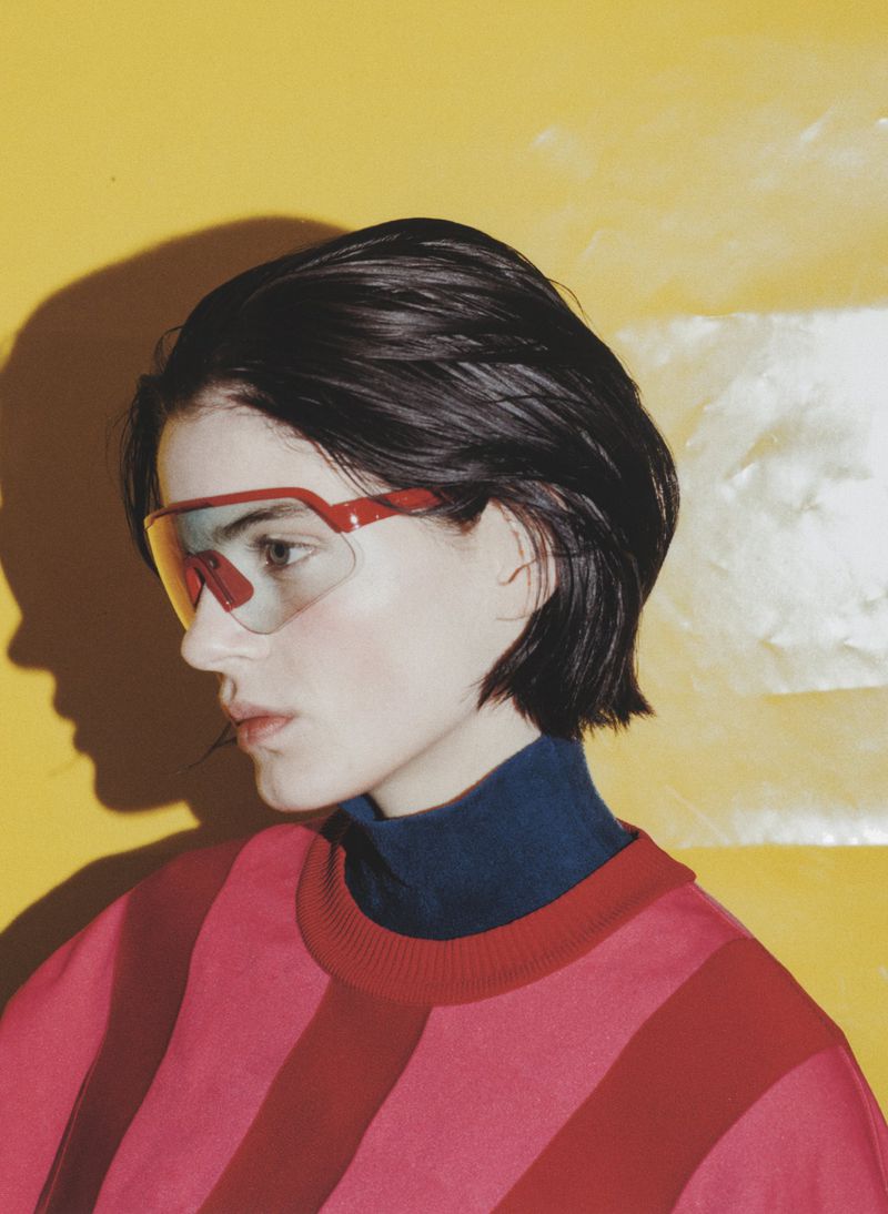 Stylish brunette woman with red-framed glasses and a pink striped sweater poses against a vibrant yellow backdrop in a studio setting.