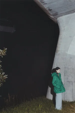 Model wearing a bright green jacket standing beneath a concrete bridge structure at night